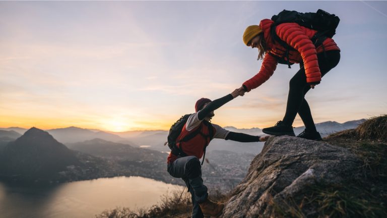 Female mountain climber helping male mountain climber reach the top of a rock with mountain landscape in distance.